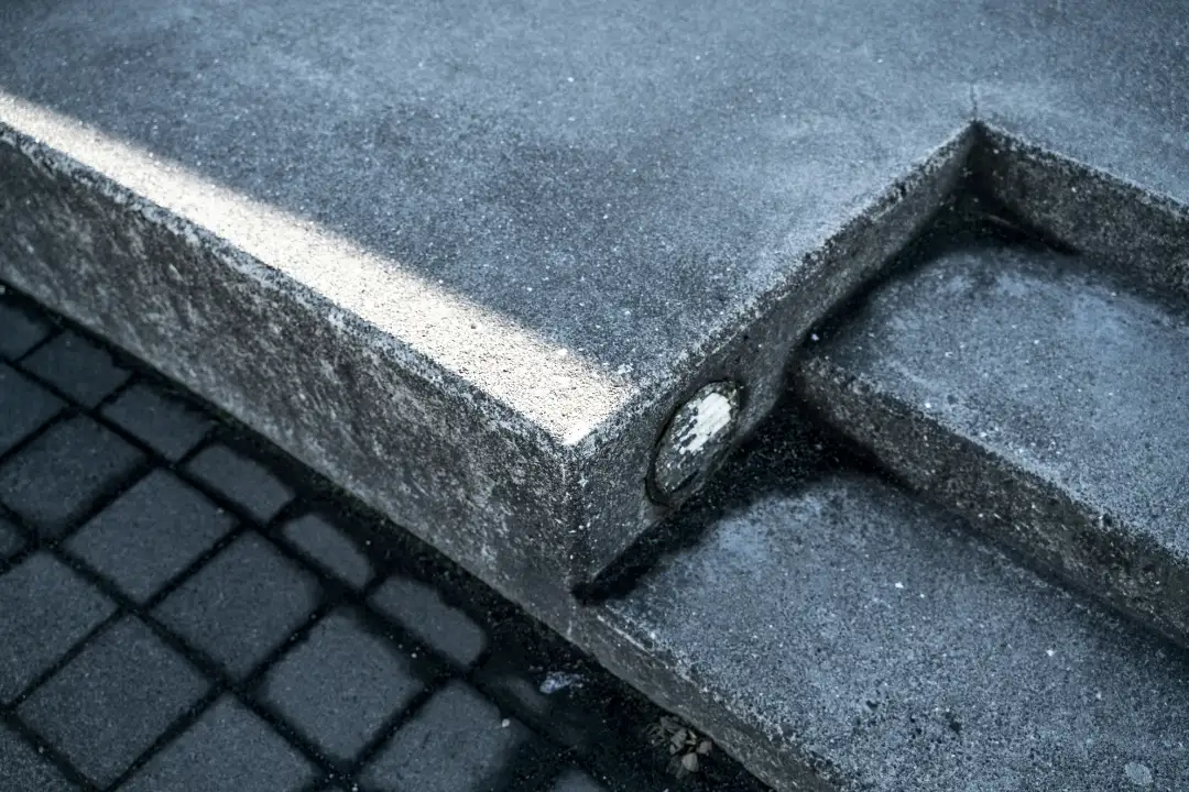 Zbliżenie na narożnik zniszczonego betonowego krawężnika, w którego boku osadzono niewielki odblaskowy metalowy znacznik; poniżej widoczna ciemna ceglana nawierzchnia – całość utrzymana w chłodnym, surowym miejskim klimacie.